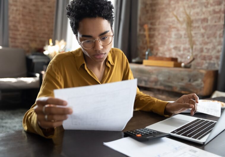 young adult looking over paperwork at a table with her laptop open