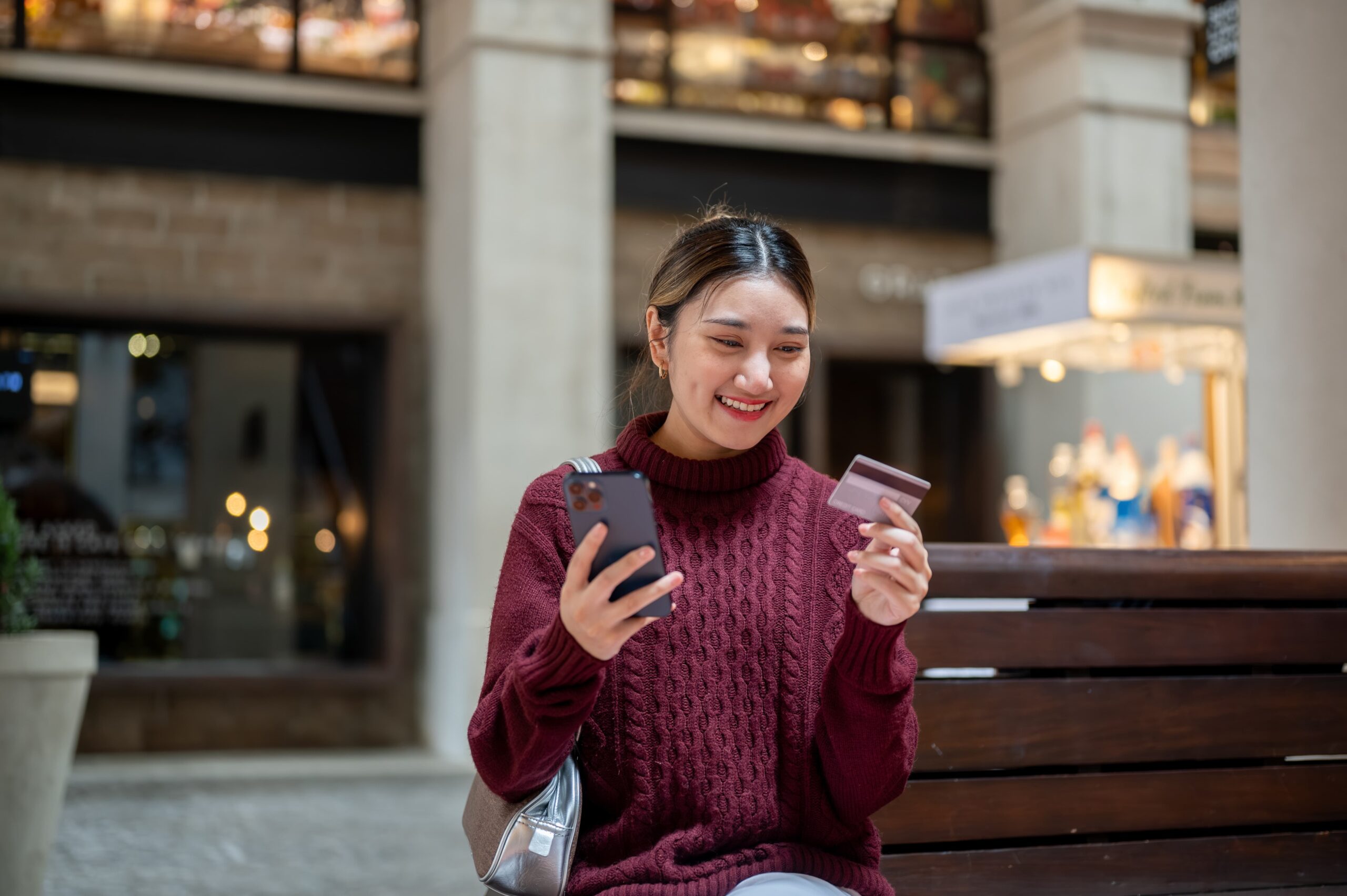 Young woman budgeting and building credic on her smartphone at a mall while holding her credit card in her other hand
